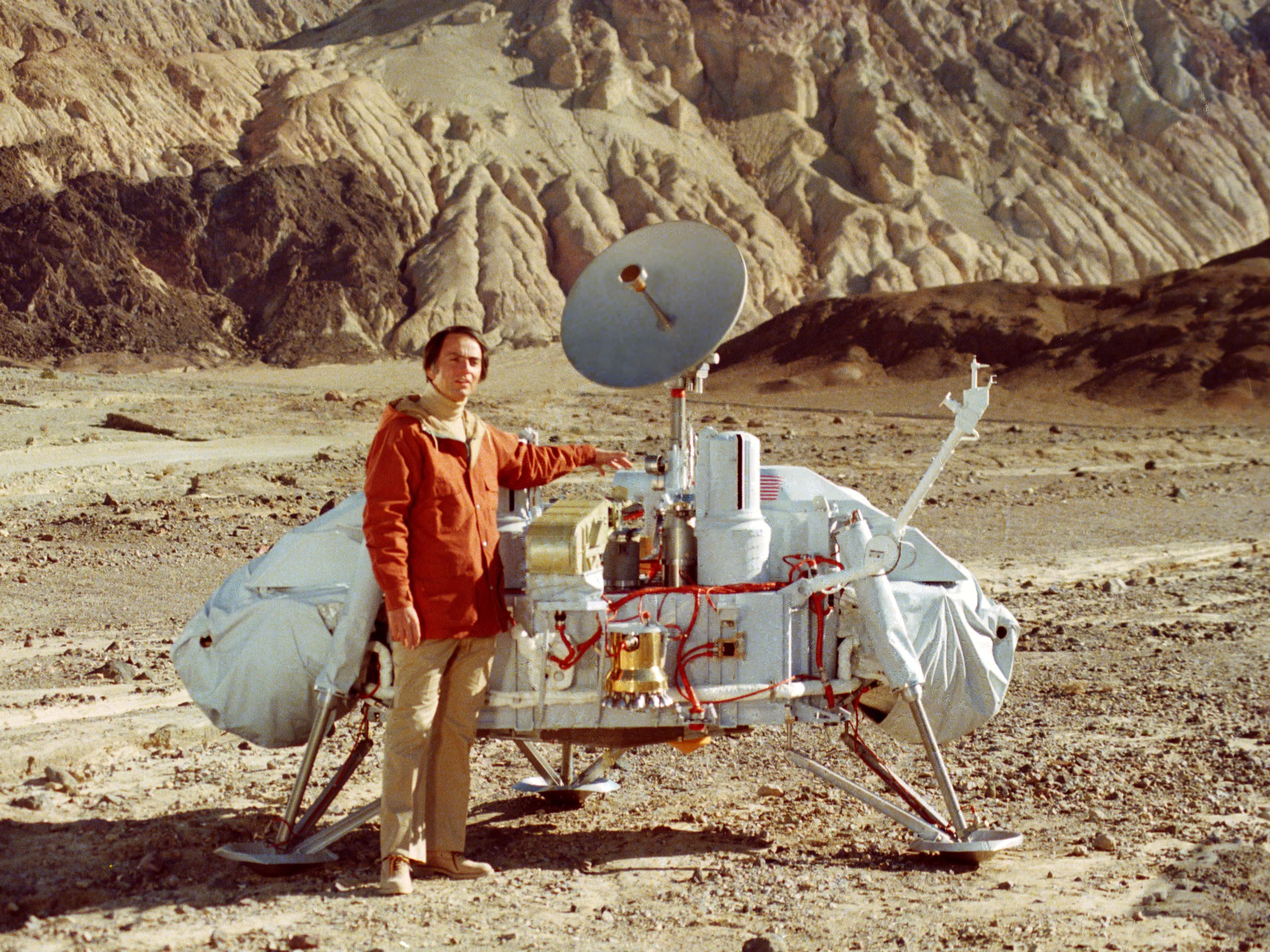 Carl Sagan posa junto a un modelo del módulo de aterrizaje Viking en un paisaje desértico.
